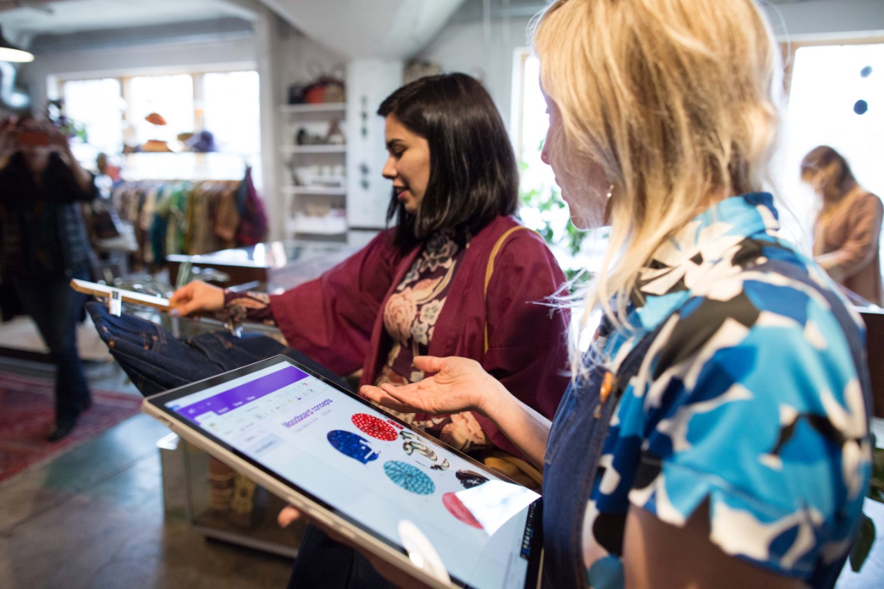 Dos mujeres interactuando en una tienda. Una sostiene un dispositivo mientras observa un catálogo digital de productos. La otra revisa una prenda de vestir. El ambiente es acogedor y está lleno de ropa y accesorios.