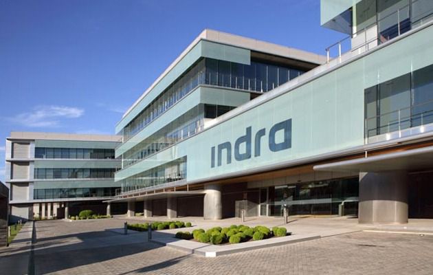 Fachada del edificio de la compañía Indra, con su logotipo visible en la parte frontal y un entorno exterior con vegetación y cielo despejado.