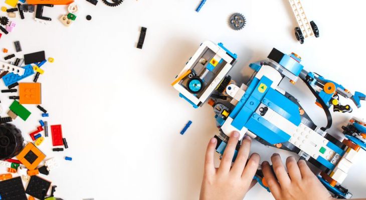 Manos de un niño ensamblando un vehículo de juguete de construcción, rodeado de piezas de lego coloridas en una mesa blanca.