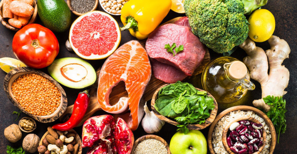 Variedad de alimentos saludables, incluyendo pescado, carne, frutas, verduras, legumbres, nueces y aceite de oliva dispuestos sobre una mesa.