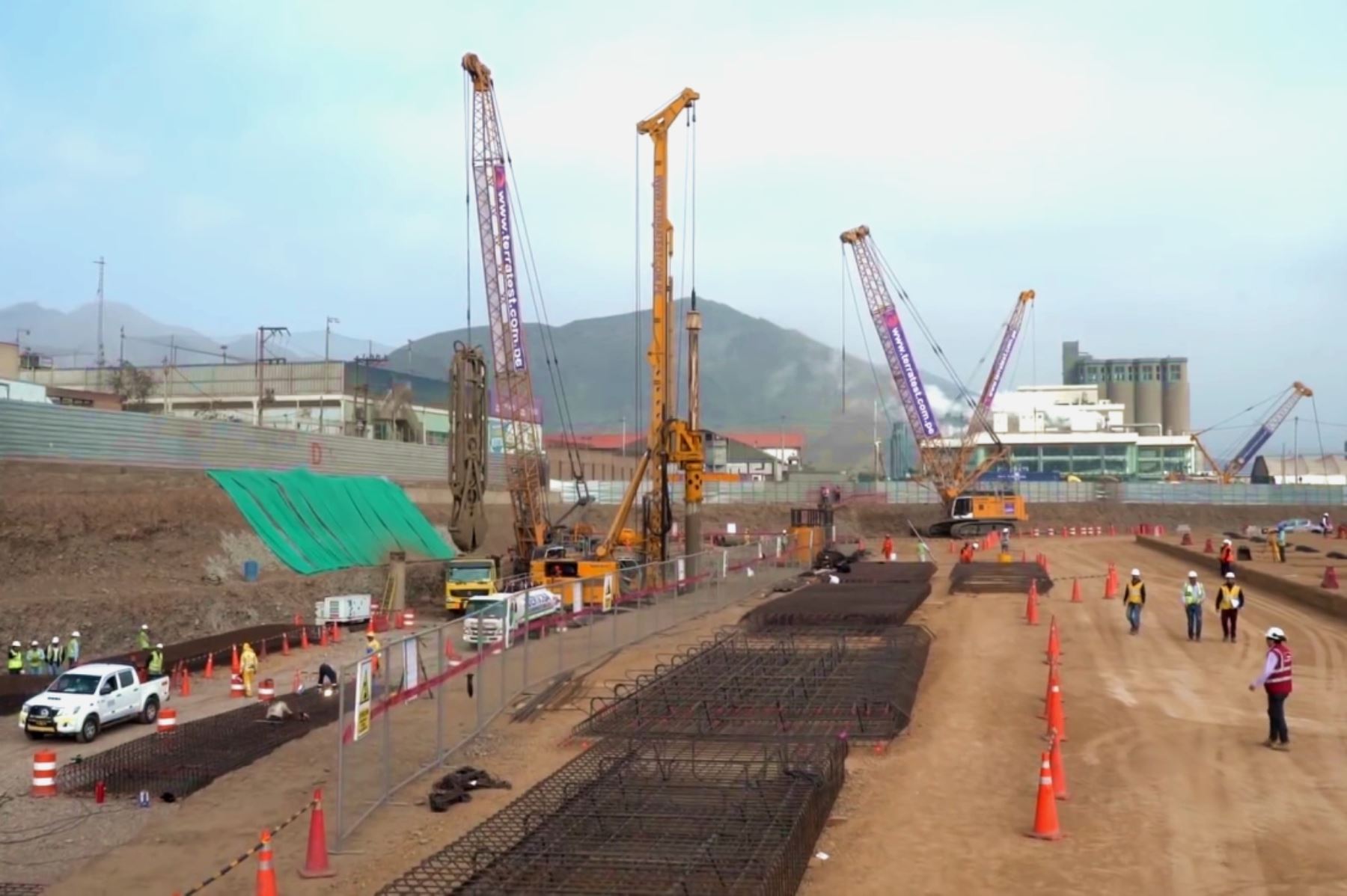 Obras de construcción en un sitio, con maquinaria pesada y trabajadores en el área. Se observa una línea de conos de tráfico y estructuras de refuerzo de acero en el suelo, así como un fondo con edificaciones y montañas.