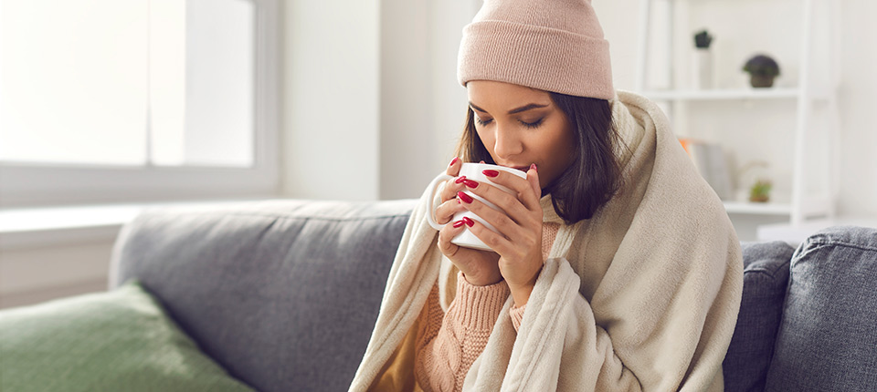 Mujer sentada en un sofá envuelta en una manta, sosteniendo una taza caliente mientras se abriga con un gorro durante el invierno.