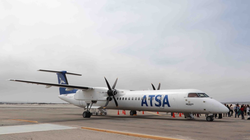 Un avión de la compañía ATSA estacionado en la pista, con fondo nublado y pasajeros formándose en la cola.