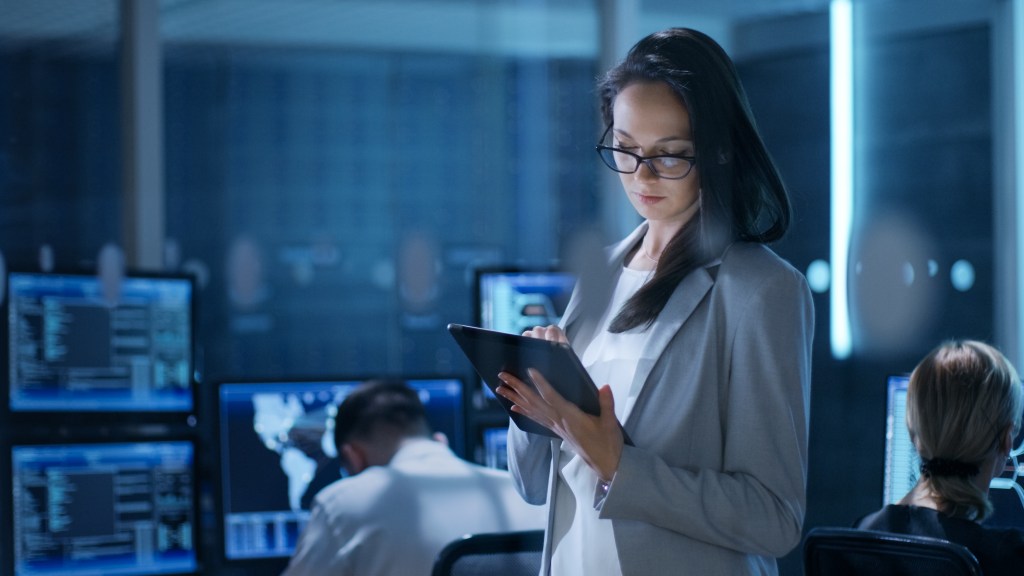 Una mujer en un entorno de oficina moderna sostiene una tableta y examina información, mientras que en segundo plano hay pantallas con datos y gráficos.