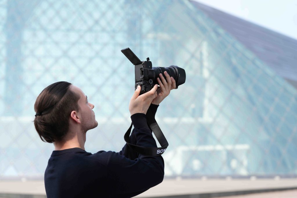 Un fotógrafo sostiene su cámara apuntando hacia arriba, con una estructura moderna de fondo.