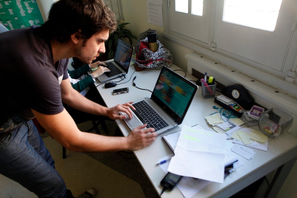 Una persona de pie interactuando con una laptop mientras otra persona trabaja en una computadora portátil en una mesa desordenada con papeles y objetos de oficina.