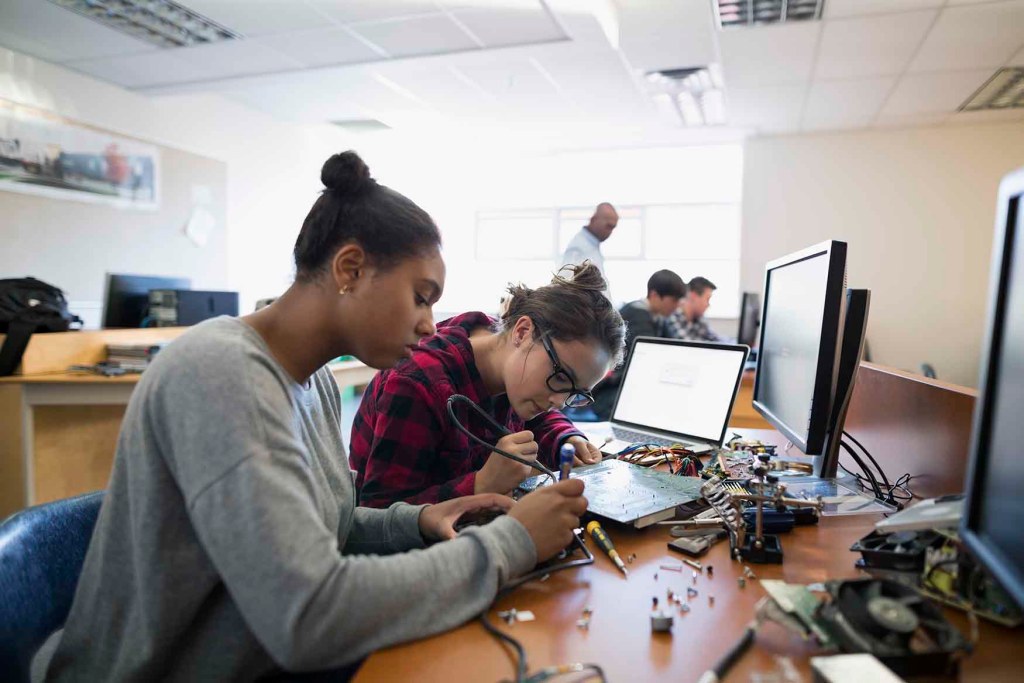 Dos mujeres jóvenes trabajando en un laboratorio, una soldando componentes electrónicos mientras la otra observa, con computadoras y herramientas de trabajo alrededor.