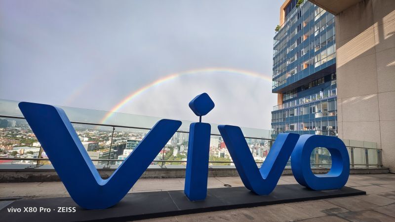 Logo de vivo en color azul en una terraza, con un arcoíris visible en el fondo y edificios modernos alrededor.