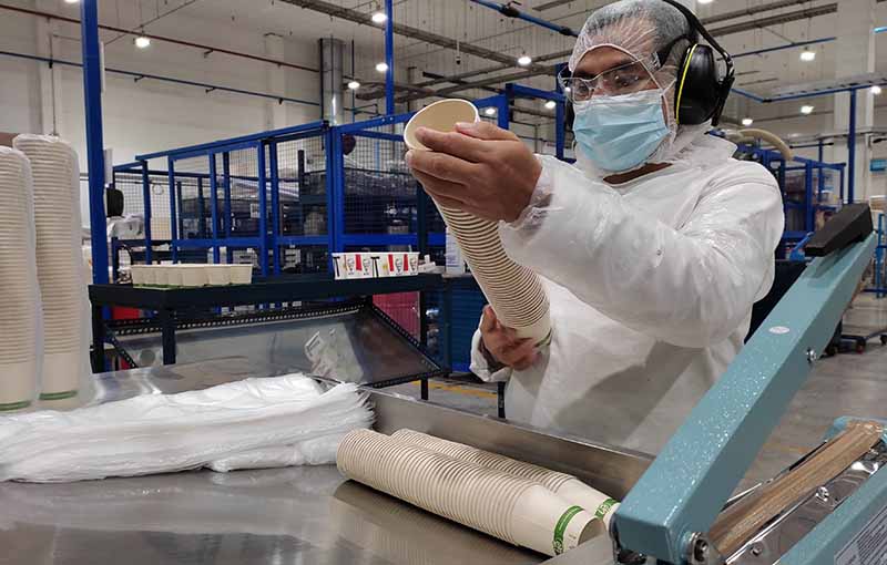 Trabajador en una fábrica inspeccionando tazas de cartón mientras lleva guantes, máscara y gorra de protección.
