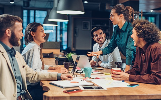 Reunión de equipo en una oficina moderna, con cinco personas sentadas alrededor de una mesa, interactuando y discutiendo proyectos.