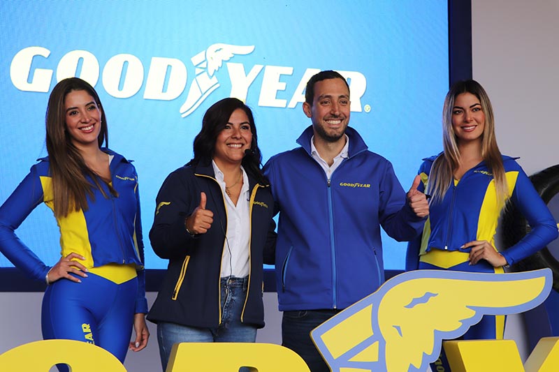 Cuatro personas posando frente a un fondo con el logo de Goodyear, dos mujeres vestidas con trajes de la marca, una mujer en chaqueta y un hombre en camisa de Goodyear, todos sonriendo y haciendo gestos de aprobación.