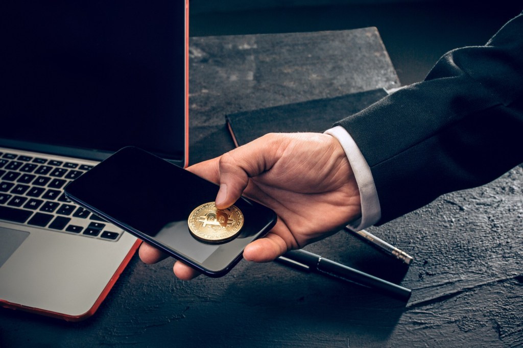 Una mano sostiene una moneda de bitcoin sobre un teléfono móvil, junto a una computadora portátil y una libreta, en un ambiente de trabajo.