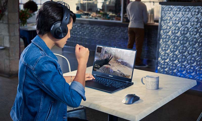 Hombre usando una laptop Lenovo en una cafetería, jugando videojuegos, con auriculares y un mouse al lado.