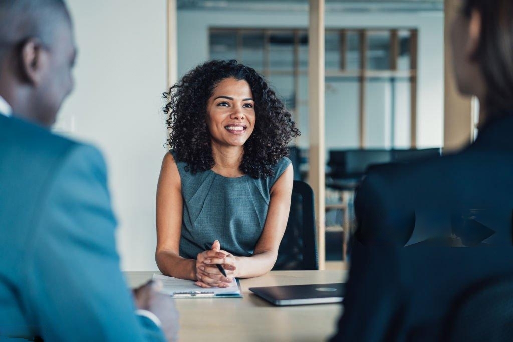 Una profesional joven con cabello rizado, sonriendo, sentada frente a dos personas, en un ambiente de oficina moderna.