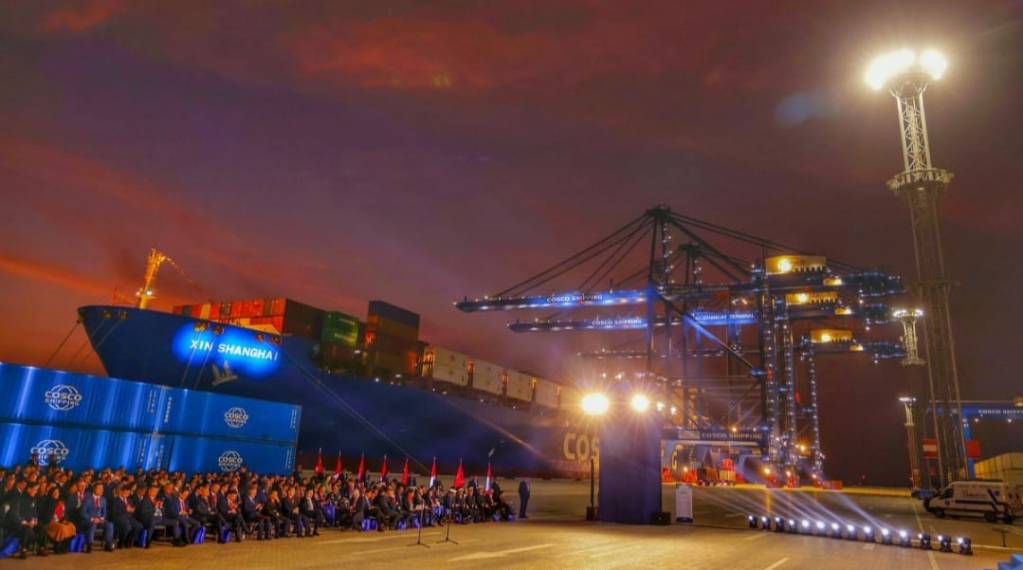 El Megapuerto de Chancay con el buque 'Xin Shanghai' atracado y un evento empresarial en primer plano, luces y una multitud de personas admirando las instalaciones del puerto al atardecer.