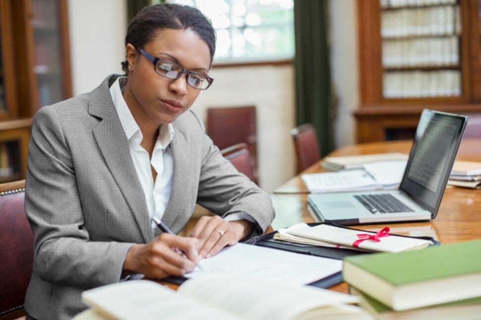 Mujer con gafas escribiendo en un cuaderno en una mesa de estudio, rodeada de libros y una computadora portátil.