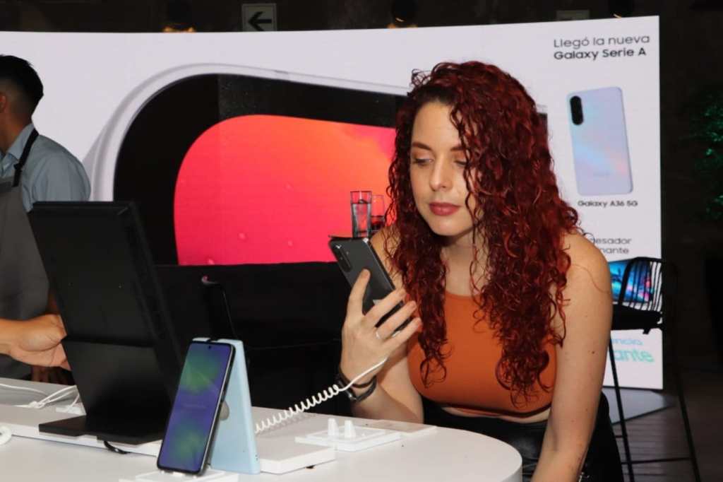 Una mujer con cabello rizado y rojizo interactúa con un smartphone en un evento de lanzamiento de celulares, con una pantalla grande de fondo que presenta el nuevo Galaxy Serie A.
