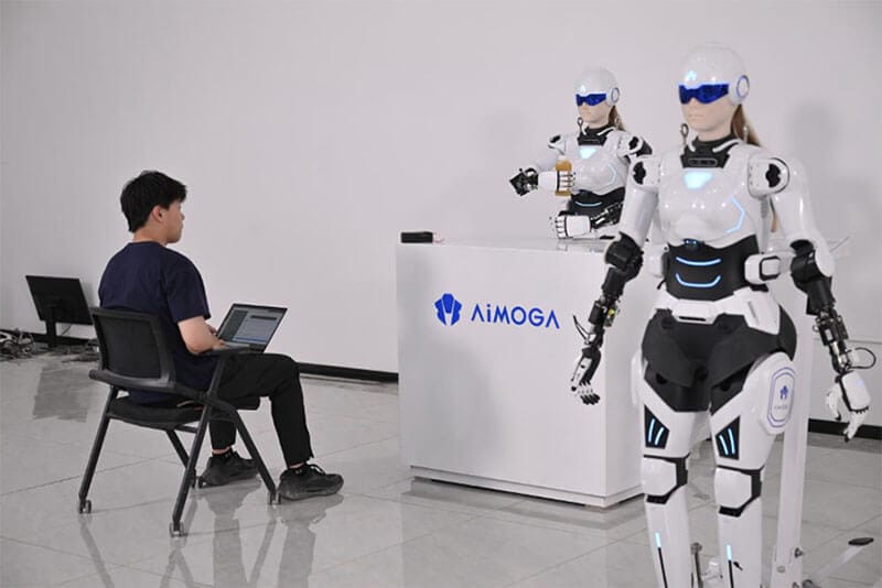 Un hombre sentado frente a un robot humanoide llamado AiMOGA, que se encuentra en una mesa con el logotipo de AiMOGA, mientras interactúa con otro robot al fondo en un ambiente moderno.