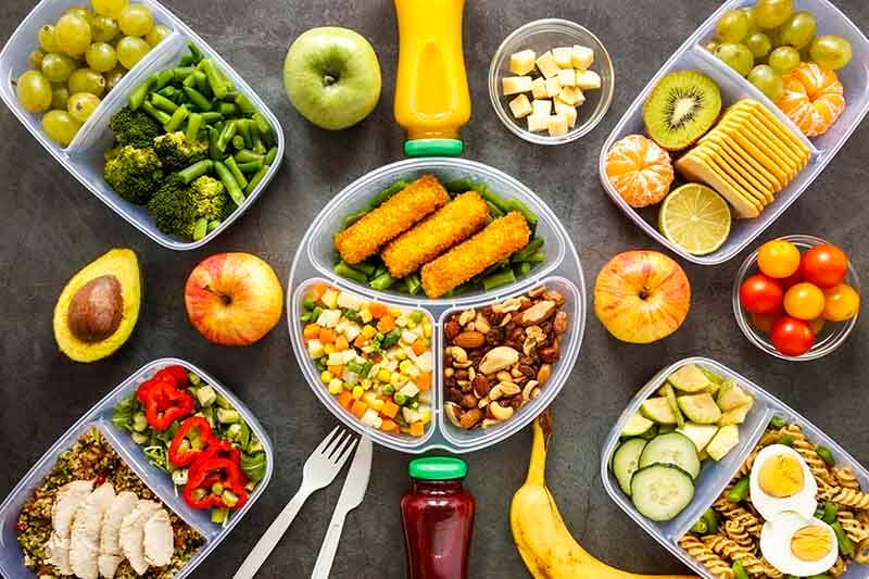 Variada selección de frutas y verduras en recipientes, incluyendo uvas, manzanas, kiwi, zanahorias, brócoli, y alimentos como pollo y nueces, sobre una superficie gris.