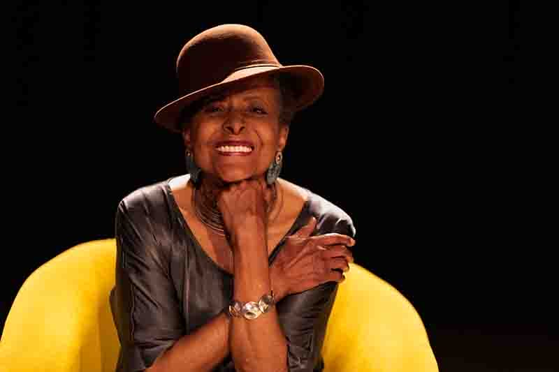 Susana Baca sonríe mientras está sentada en una silla amarilla, vistiendo una blusa gris y un sombrero marrón, en un ambiente oscuro.