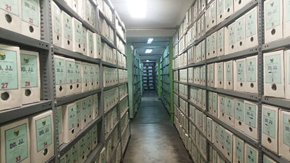 Pasillo de archivos organizados en cajas etiquetadas, iluminado por luces fluorescentes, mostrando un entorno de almacenamiento de documentos.