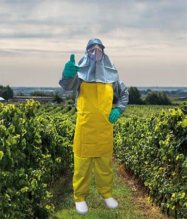 Trabajador agrícola en un campo con equipo de protección personal, incluyendo guantes, traje amarillo y una máscara, haciendo un gesto positivo con el pulgar hacia arriba.