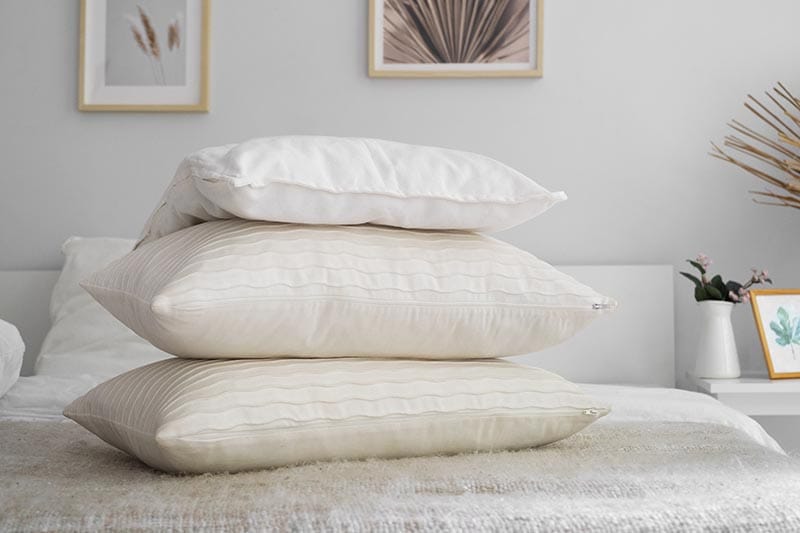 Una pila de tres almohadas de diferentes texturas y colores sobre una cama, con un ambiente decorativo moderno y minimalista en el fondo.