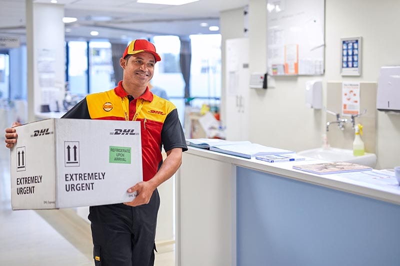 Un mensajero de DHL, vestido con un uniforme rojo y amarillo, sostiene una caja marcada como 'EXTRAMAMENTE URGENTE' en un entorno de oficina médica.