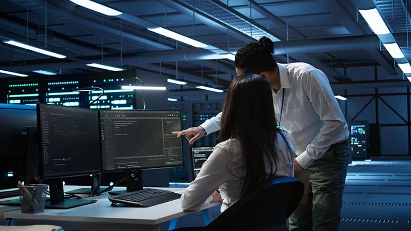 Dos profesionales de ciberseguridad colaborando frente a monitores en un entorno de oficina moderna, analizando información en pantallas de computadora.