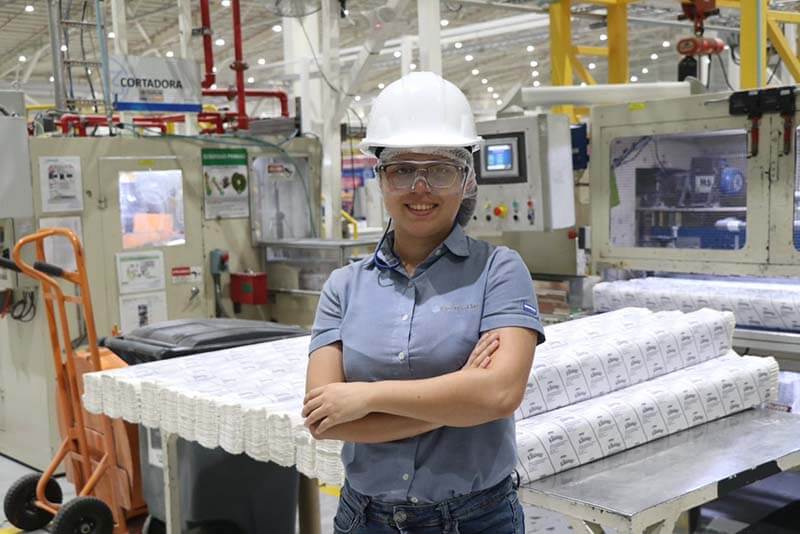 Mujer con casco y gafas de seguridad en una planta de producción, sonriendo mientras se apoya en una mesa con productos empaquetados.