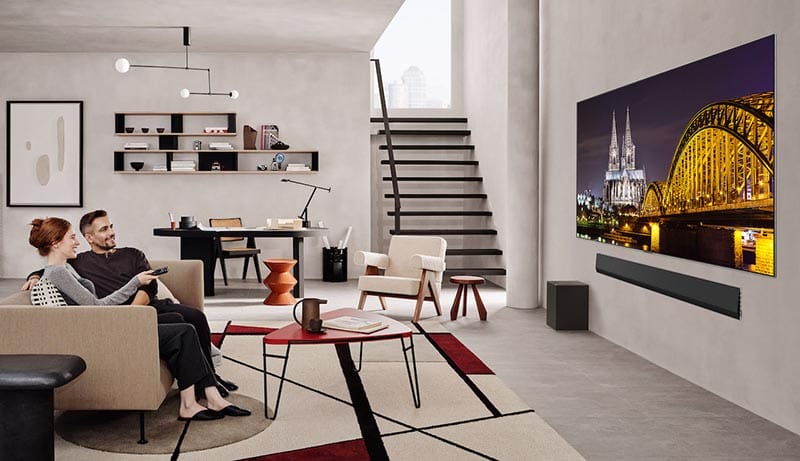 Una pareja sentada en un sofá moderno en una sala de estar, mirando un televisor grande que muestra una imagen de un paisaje urbano iluminado por la noche. La decoración incluye una mesa de café y estantes con objetos decorativos.