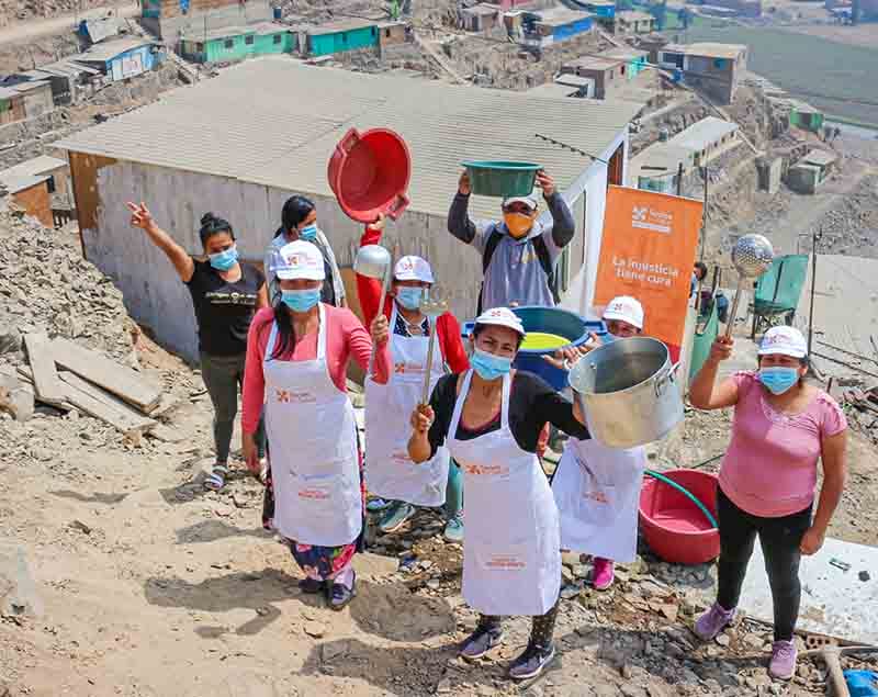 Grupo de mujeres sonrientes al aire libre, usando mascarillas y delantales, sosteniendo recipientes de cocina en un entorno de construcción en Carabayllo.