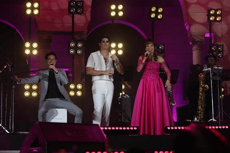 Tres artistas en un escenario iluminado, dos cantantes y un músico, durante un evento de karaoke organizado por Mallplaza en Perú. Uno de los cantantes viste una camisa blanca y el otro una vestido rosa, mientras que el músico tocando un cajón está a su lado.