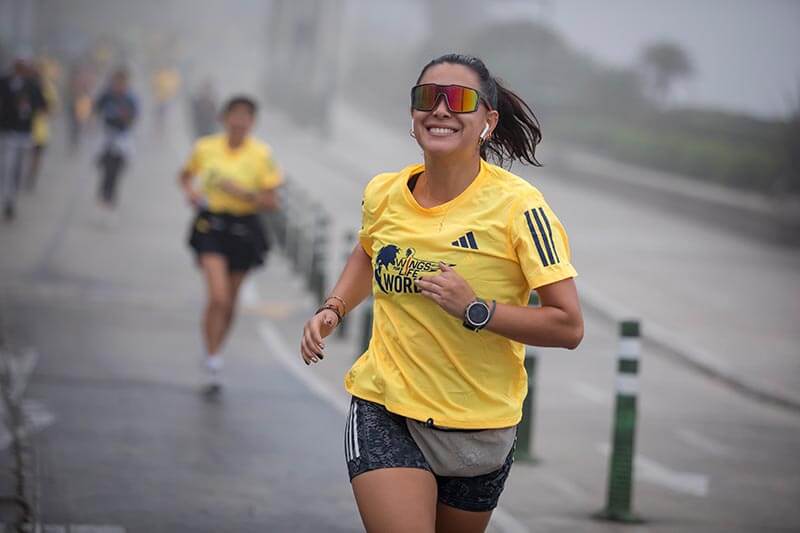Una mujer sonriente corre durante la Wings for Life World Run, vistiendo una camiseta amarilla con el logo del evento y gafas de sol. El ambiente es nublado y hay otros corredores al fondo.