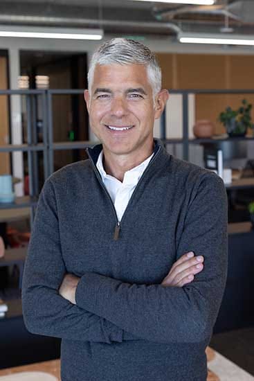 Ejecutivo brasileño sonriendo con los brazos cruzados, vistiendo un suéter oscuro, en un entorno de oficina moderna.