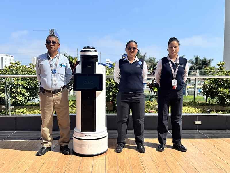 Tres personas de pie junto a un robot de patrullaje, en un entorno al aire libre con vegetación y un cielo despejado.