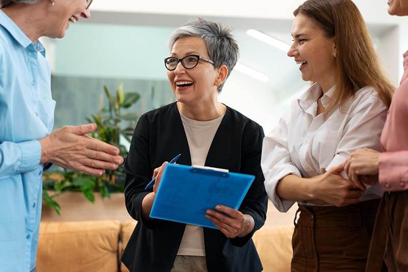 Un grupo de cuatro profesionales conversando en un ambiente informal, con una mujer de cabello corto y canoso sosteniendo una carpeta azul mientras sonríe.