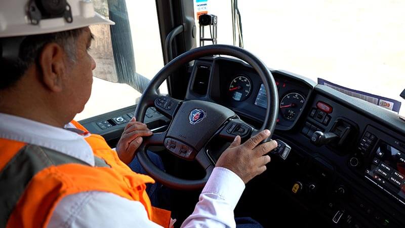 Conductor de camión usando un casco y chaleco de seguridad, asistiendo al panel de control del vehículo en un ambiente de trabajo.