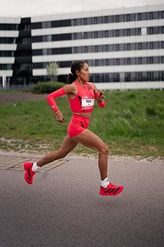 Thalía Valdivia, atleta peruana, corre con determinación en una competencia, vistiendo un atuendo deportivo rojo y zapatillas adidas, con un moderno edificio de fondo.