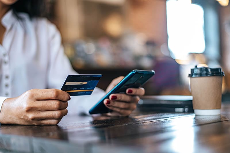 Una persona sosteniendo una tarjeta de crédito en su mano izquierda, mientras usa un teléfono móvil con la mano derecha, sobre una mesa de madera. Al fondo, se puede ver una taza de café y un ambiente de cafetería.