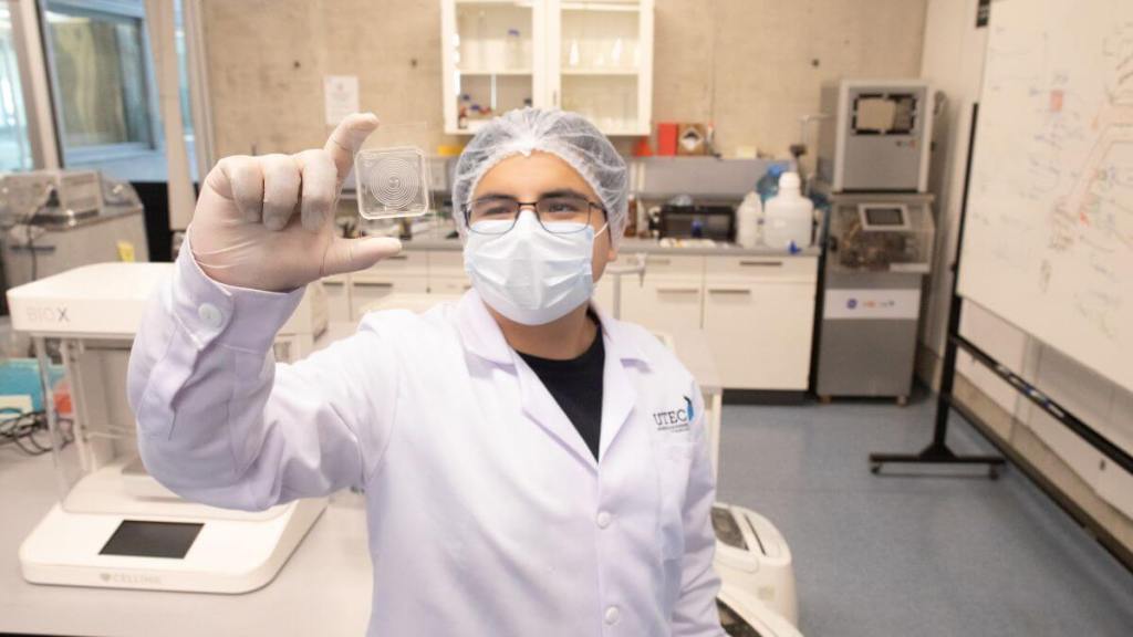 Un científico de la UTEC sostiene un pequeño dispositivo innovador, el 'Chip de la esperanza', en un laboratorio, con equipo de laboratorio y herramientas visibles al fondo.