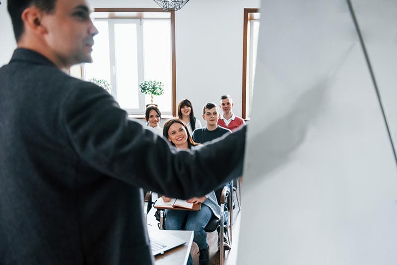 Una clase en un aula moderna donde un instructor está presentando a un grupo de estudiantes interesados, todos sonriendo y prestando atención.