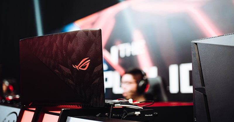 Laptop gamer ROG en un entorno de competición, resaltando su diseño y logo, con jugadores indistinguibles en el fondo.