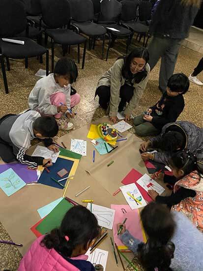 Grupo de niños y una adulta participando en actividades de arte, utilizando papel de colores y herramientas de dibujo en un ambiente de taller.