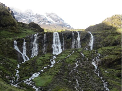 Cascadas de agua fluyendo por un paisaje montañoso verde, con picos nevados al fondo.