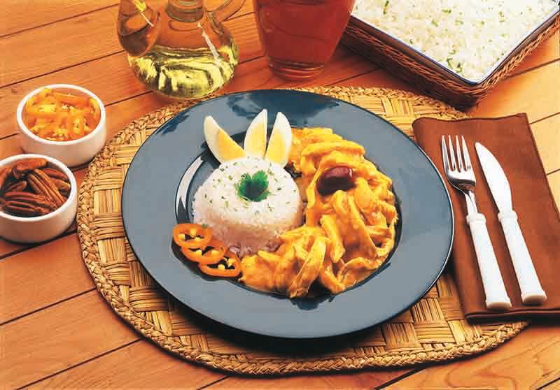 Un plato de ají de gallina servido con arroz blanco, acompañado de rodajas de limón, ají y aceitunas, sobre un fondo de mesa de madera.