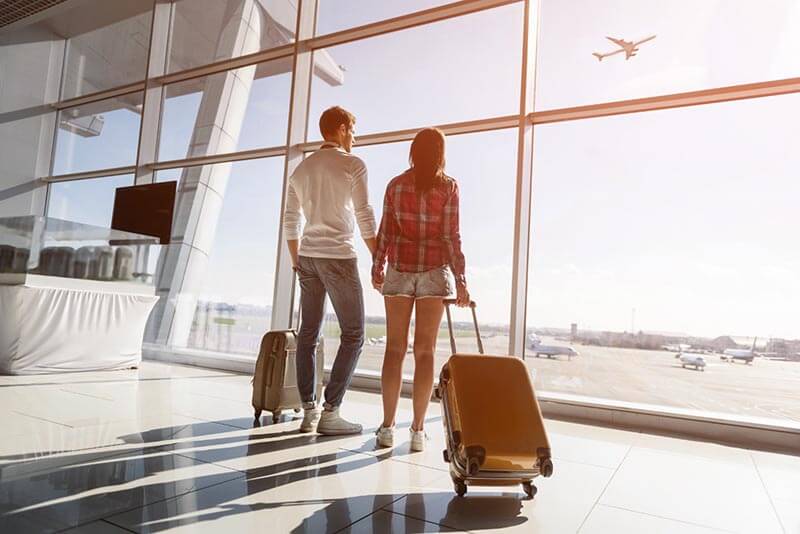 Pareja de viajeros en un aeropuerto mirando hacia el exterior mientras sostienen maletas.
