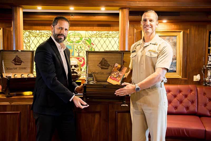 Dos hombres posan con una caja de madera emblemática y productos de cerveza Cusqueña, en un ambiente elegante a bordo de un barco escuela.