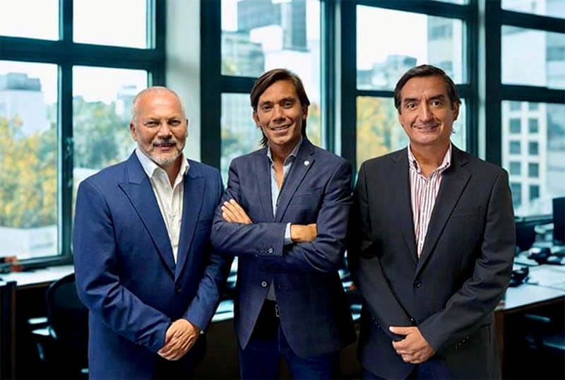 Tres hombres posando juntos en un entorno de oficina con ventanas grandes al fondo.