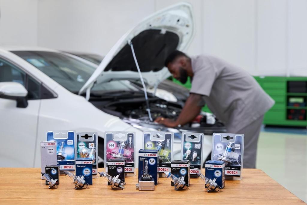 Un mecánico trabaja en el motor de un automóvil blanco, mientras en primer plano se muestran varias cajas de luces de automóviles Bosch sobre una mesa.